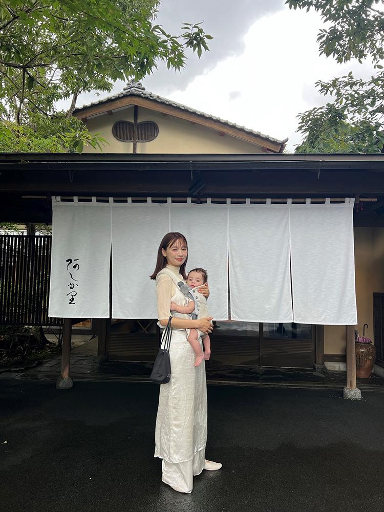 Woman in a white dress holding a baby, standing in front of a traditional Japanese building with white curtains.