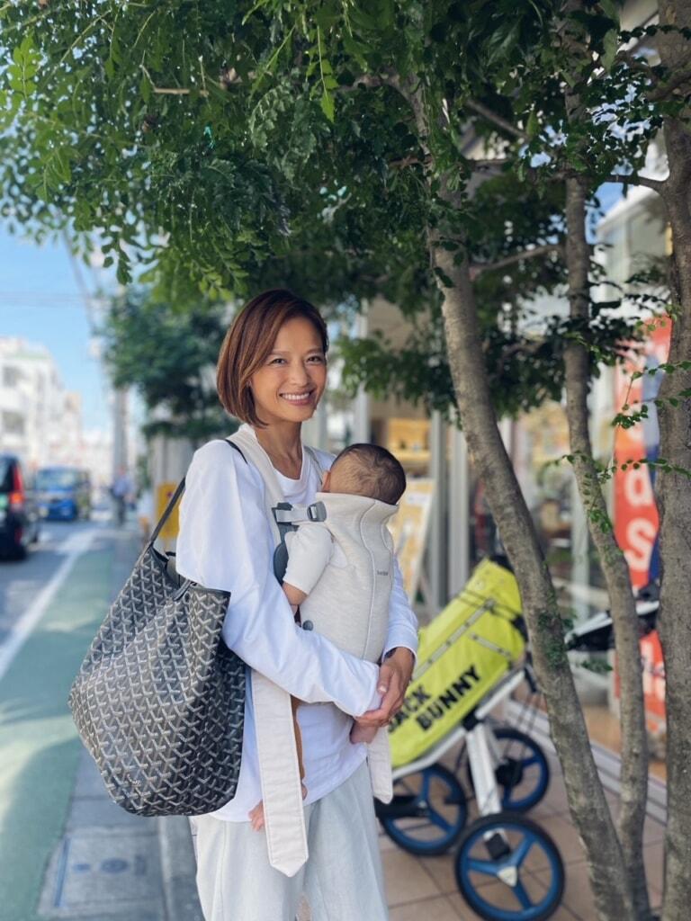 A woman smiles while holding a baby in a carrier, standing on a tree-lined sidewalk. She carries a large patterned bag over her shoulder.