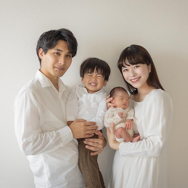A smiling family of four poses together against a plain background. The parents hold a young child and a newborn baby, all dressed in light-colored clothing.