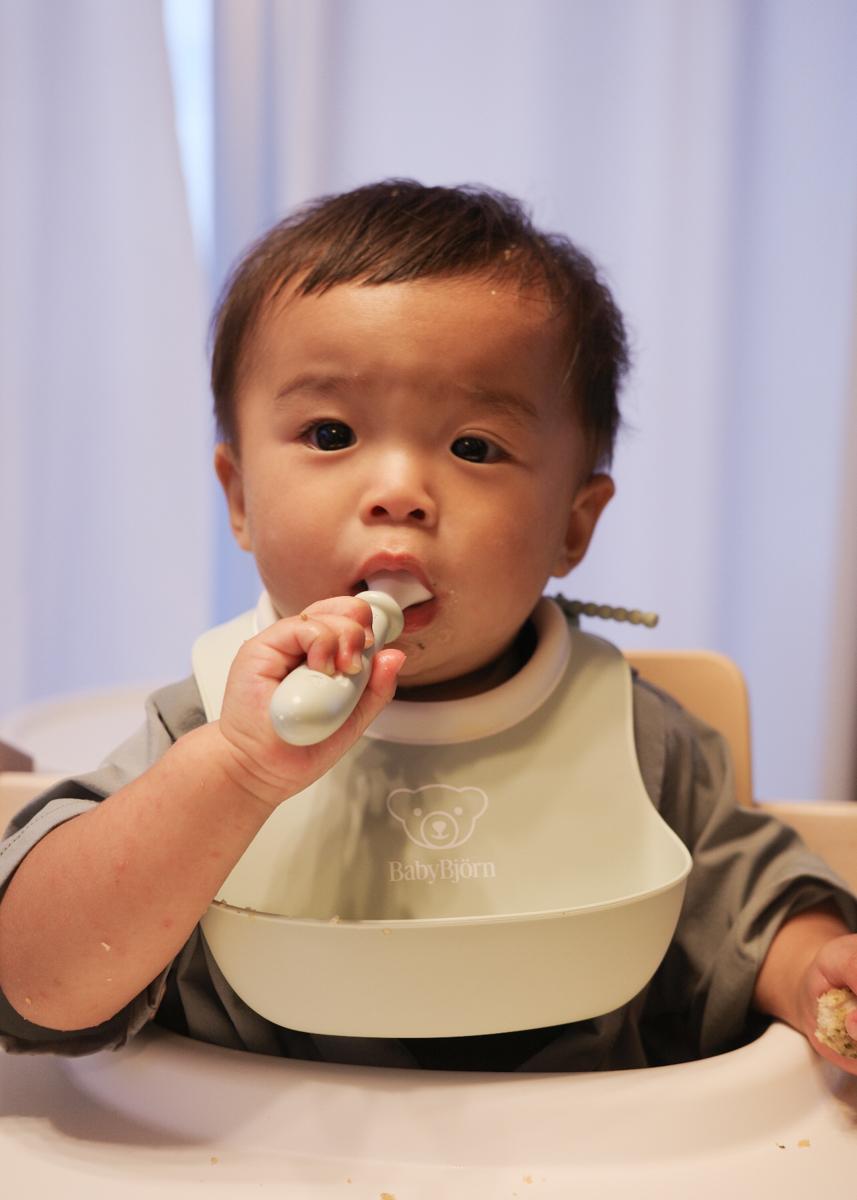A baby in a high chair wearing a bib, holding a spoon to their mouth, with a focused expression.