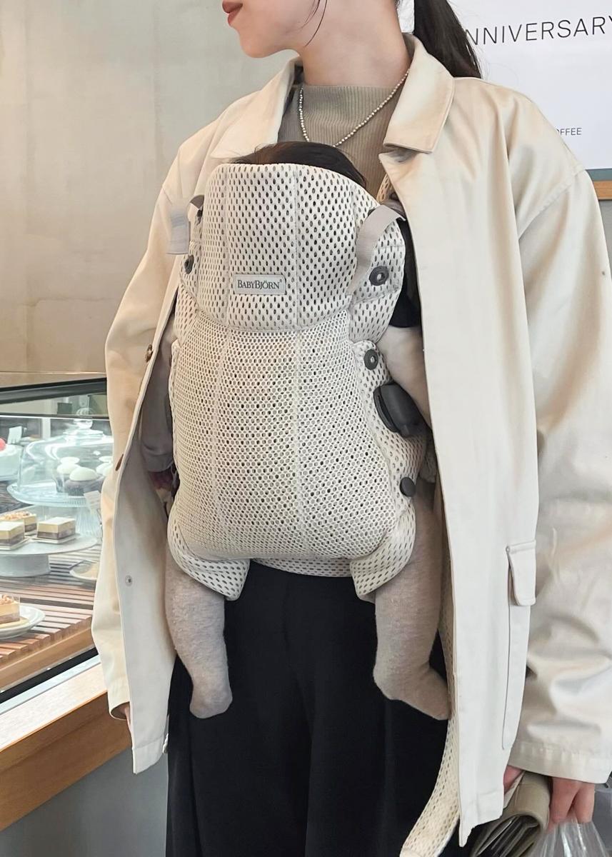 Person wearing a beige jacket carrying a baby in a gray mesh baby carrier, standing near a pastry display.