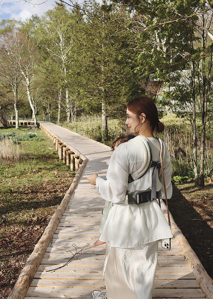 A woman in a white outfit walks on a wooden path through a lush, green forest, carrying a baby in a front carrier.