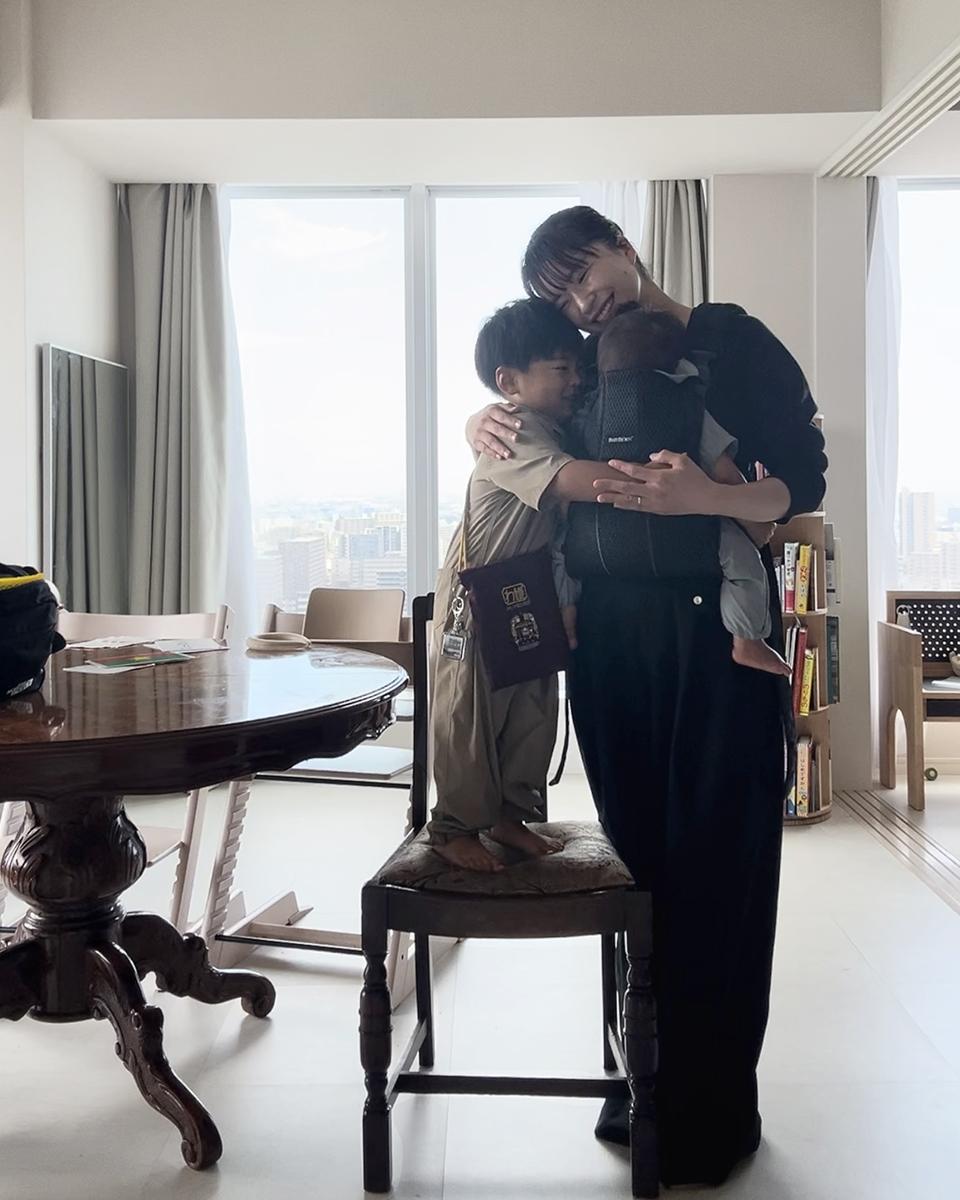 A person embraces two children in a bright room, one child standing on a chair and the other in a baby carrier.
