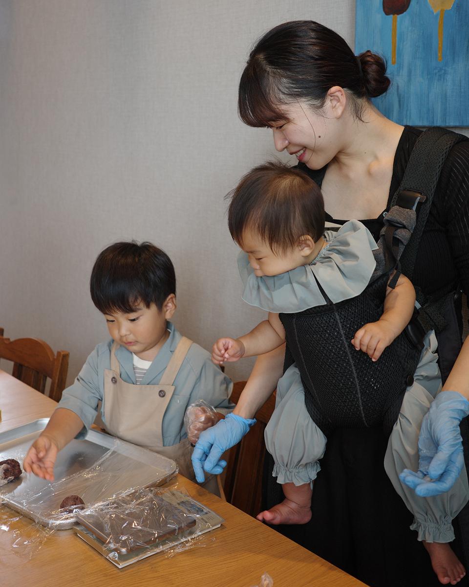 A woman with a baby in a carrier helps a young child prepare food at a table, with trays and ingredients in front of them.