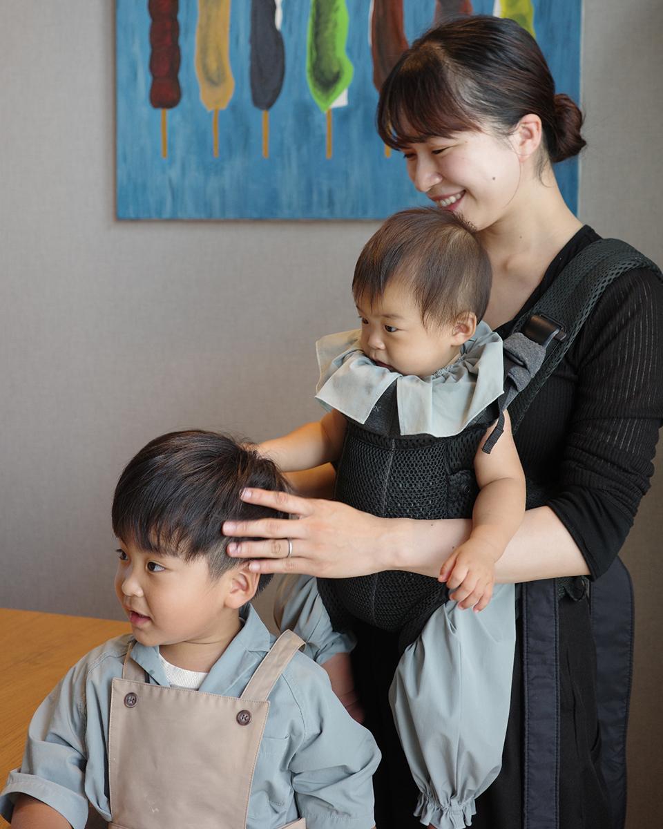 A smiling woman holds a baby in a carrier, gently touching a young boy's hair. A colorful painting is in the background.
