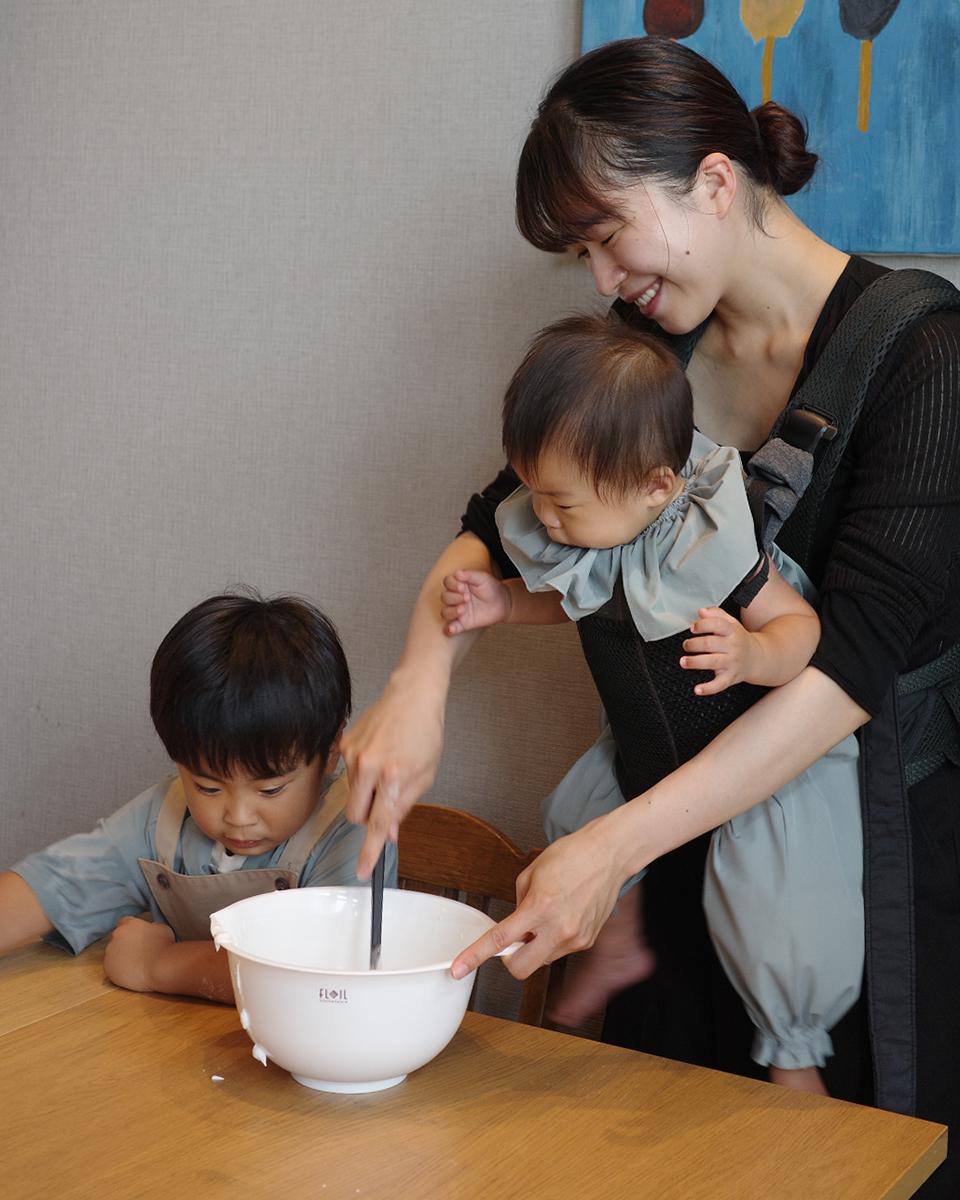 A mother helps her young son mix ingredients in a bowl, while carrying a baby in a sling. They are all wearing matching outfits.